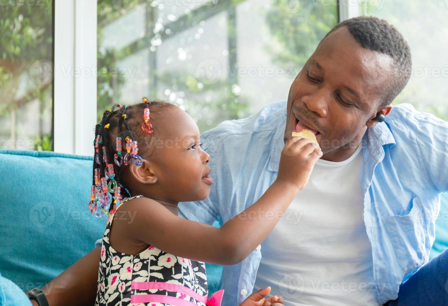 schattig klein meisje kijkt naar haar vader die een snack eet, vrolijke Afro-Amerikaanse vader en dochter die in de woonkamer spelen, geluksfamilieconcepten foto