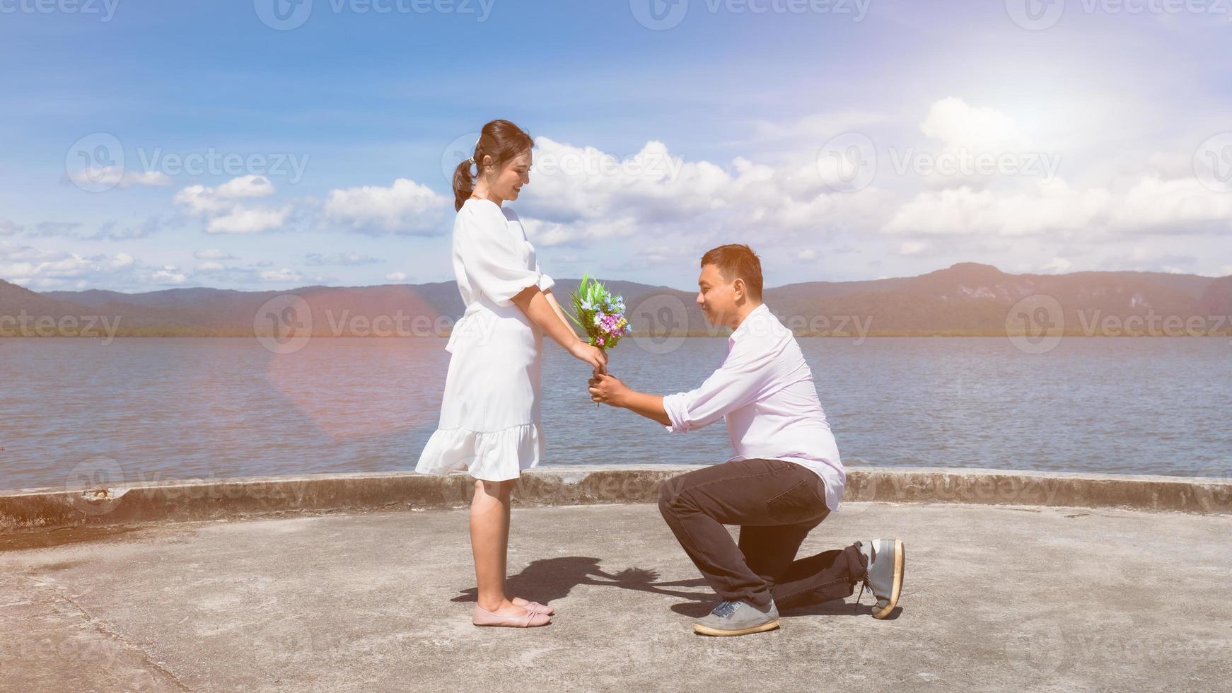 een Aziatische man knielde neer om een boeket bloemen te geven aan een Aziatische vrouw in het wit gekleed in de zeehaven. om liefde uit te drukken en voor te stellen in de zon, heldere luchten, sterke wind. foto