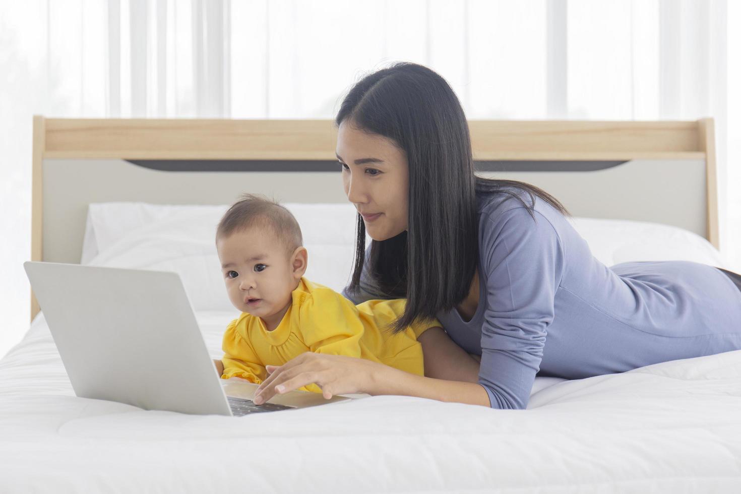 een aziatische moeder werkt op een laptop in bed en met haar baby naast het concept van werk thuis. foto