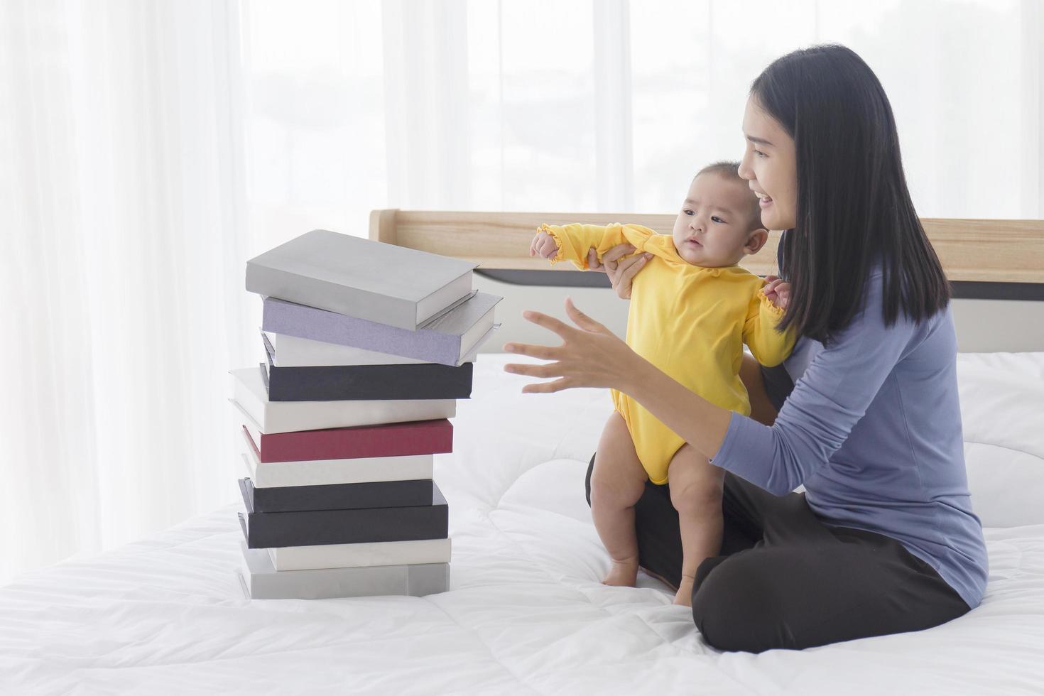 een Aziatische moeder hielp haar baby om op te staan. en er lag een stapel boeken naast haar op het bed. foto