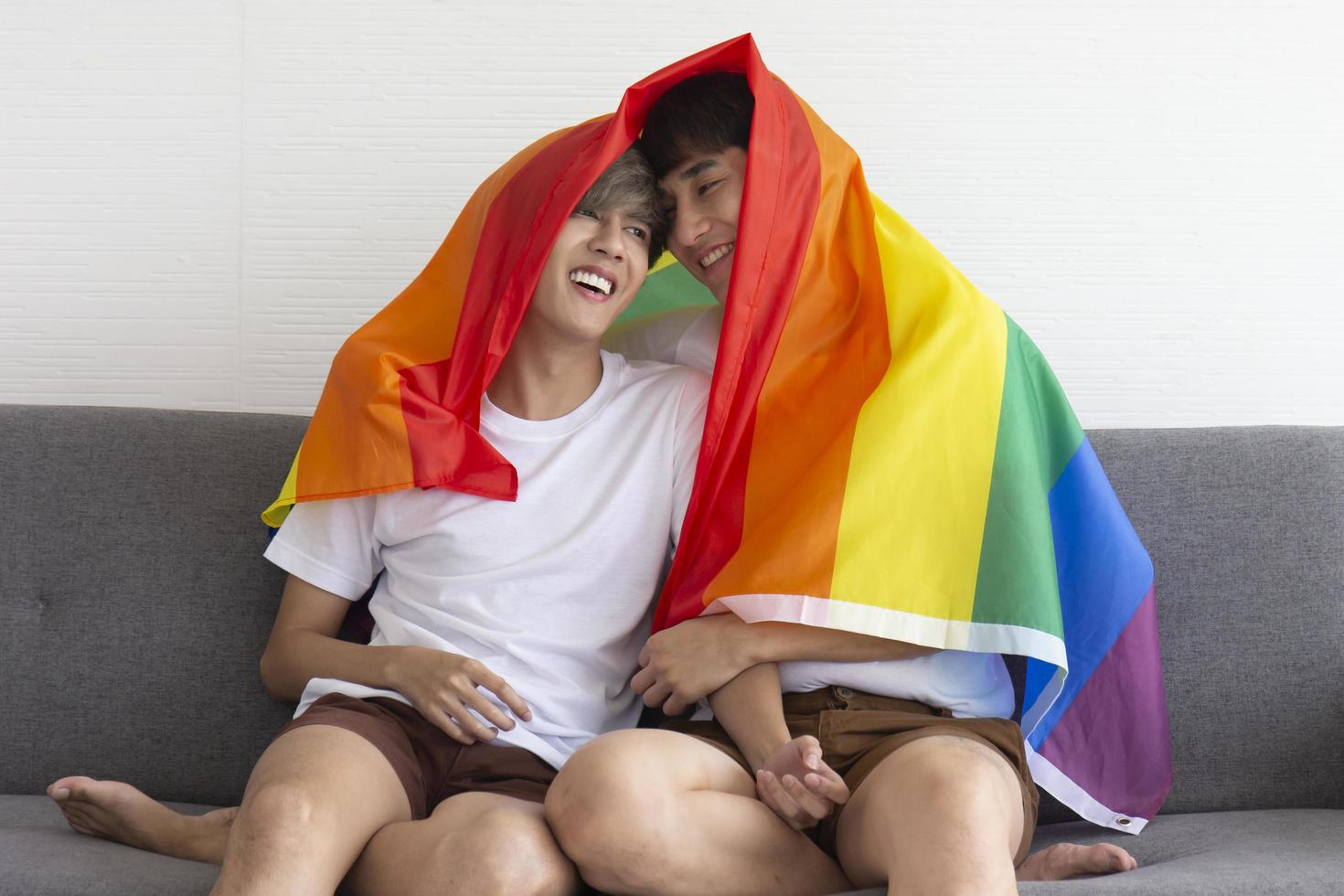 een mannelijk stel met een aziatische man zittend op een bank, met een veelkleurige vlag met tekens van lhbt op hun hoofd, openlijk homoseksuele mannen die lhbt-ideeën accepteren. foto