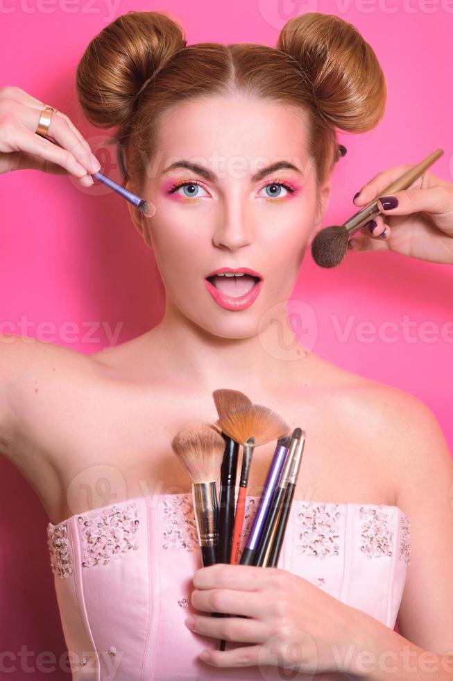 aantrekkelijke blonde vrouw met kleurrijke make-up met cosmetische penselen en schaduwen in haar en make-up master's hands foto