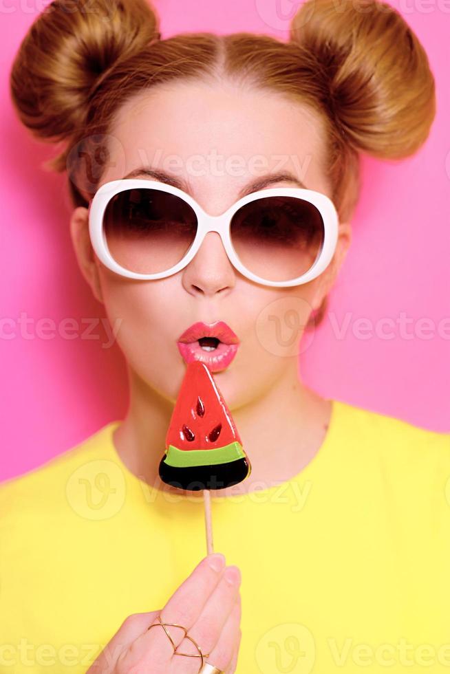 portret van stijlvolle schattige jonge blonde vrouw in zonnebril met kleurrijke watermeloen lolly op roze achtergrond foto