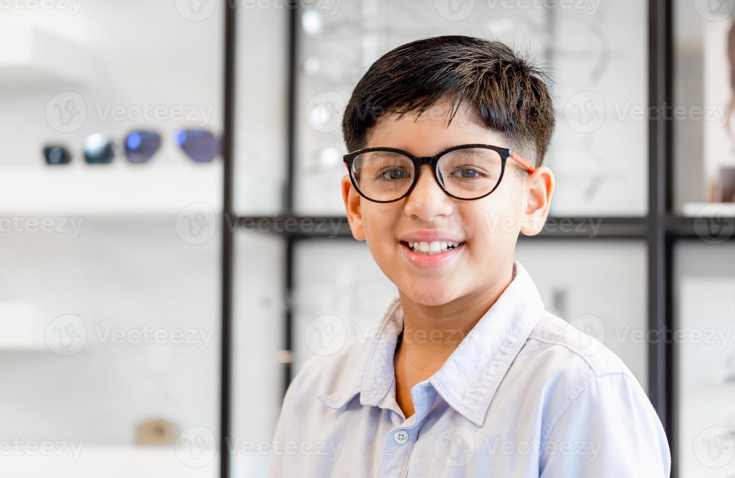 lachende indisch-thaise jongen die een bril kiest in de optiekwinkel, portret van een kind met een gemengd ras etniciteit dat een bril draagt in de optische winkel foto