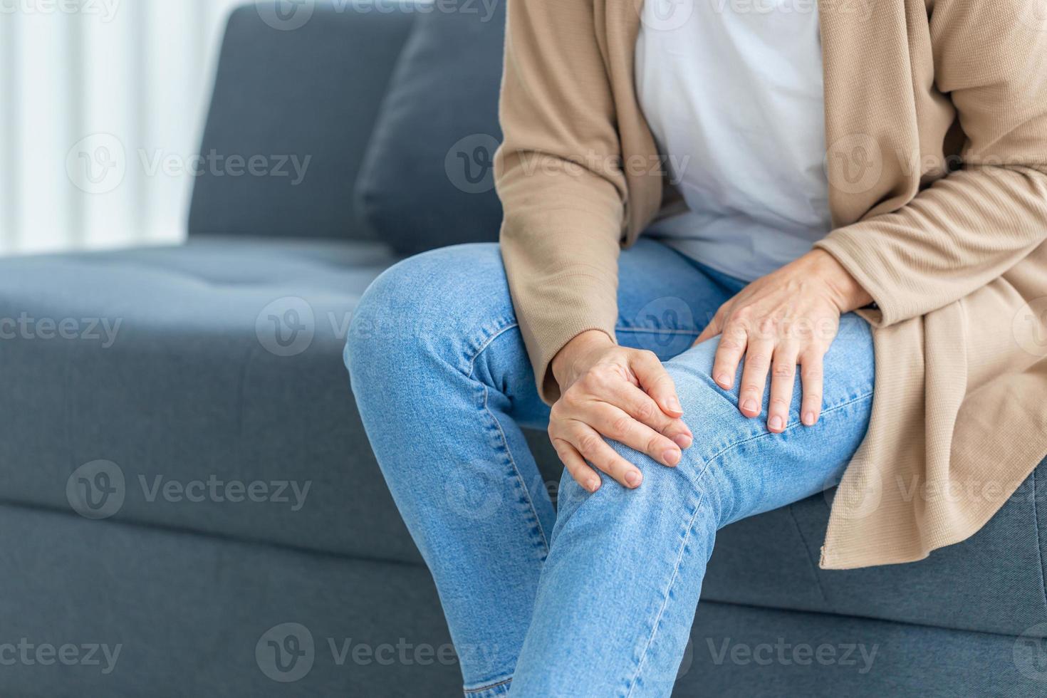 vrouw lijdt aan kniepijn zittend op de bank in de woonkamer, volwassen vrouw lijdt aan kniepijn terwijl ze op de bank zit foto