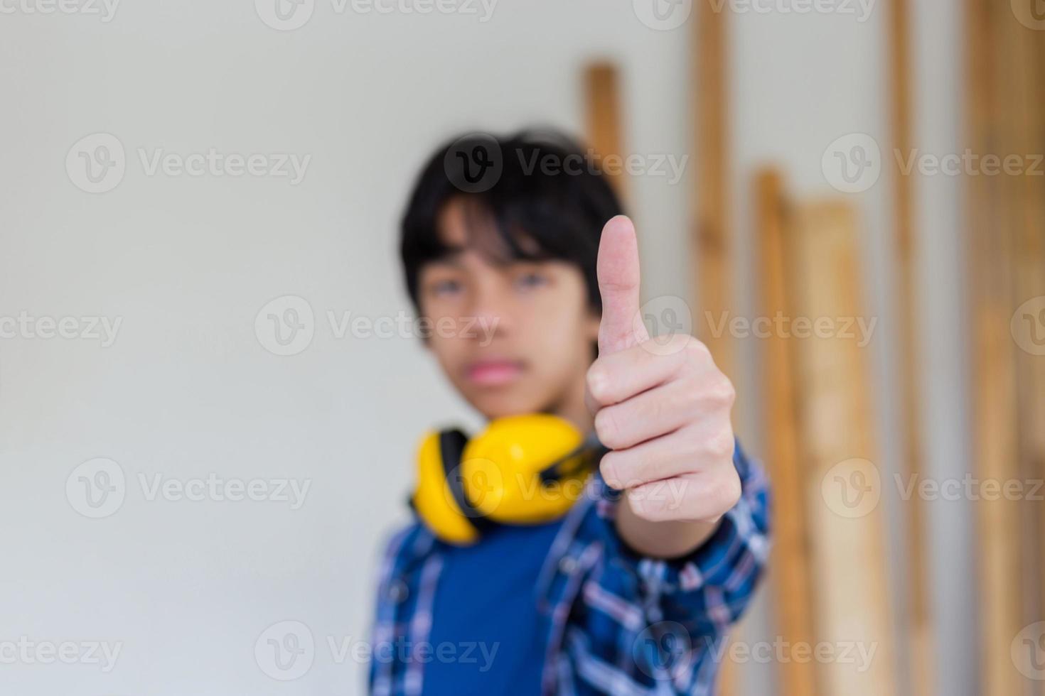 jonge jongen staat met duimen omhoog als teken van succes in een timmerwerkplaats. kind met oorkappen met ruisonderdrukking leren in de ambachtswerkplaats foto