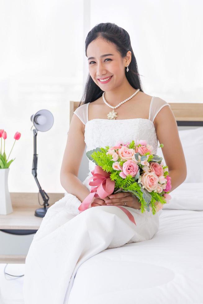 een aziatische bruid in een trouwjurk zit fel glimlachend op het bed in haar hand met een prachtig boeket bloemen. foto