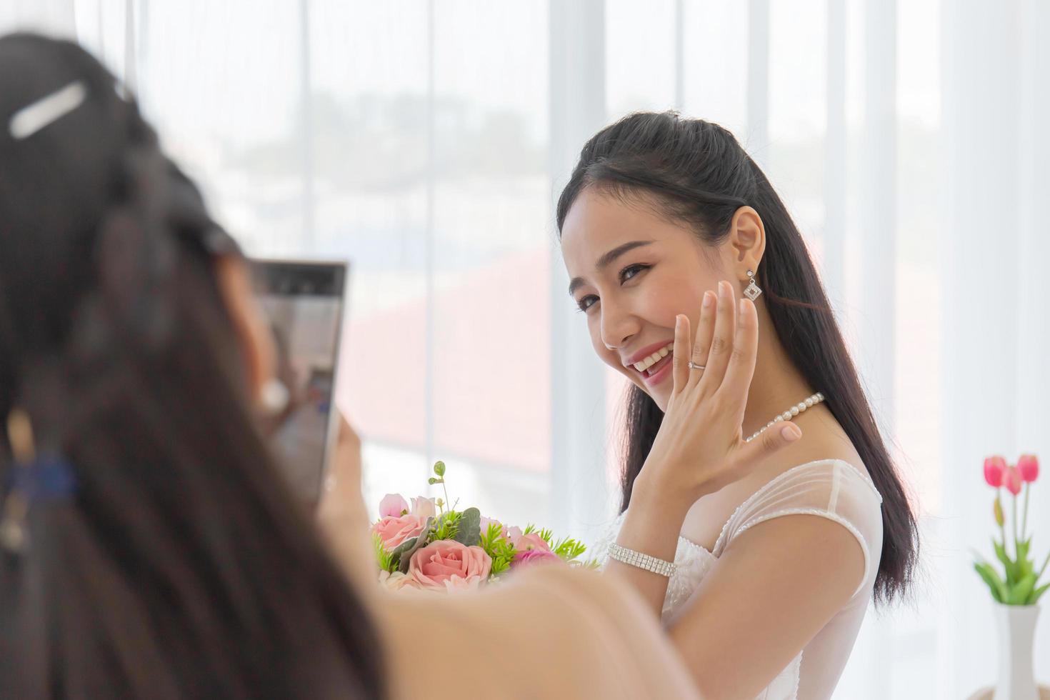 de vriendin van de bruid gebruikt een smartphone om foto's te maken van een Aziatische bruid in een trouwjurk die haar linker diamanten ring laat zien in de kleedkamer met een schattige, heldere glimlach. foto