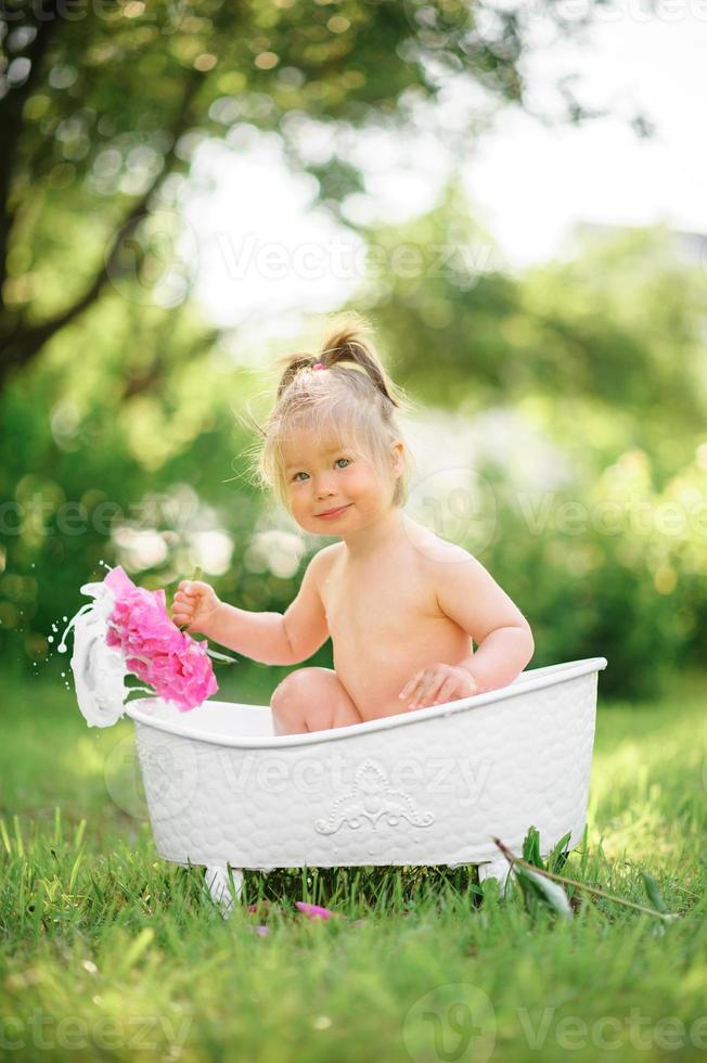 gelukkig peutermeisje neemt een melkbad met bloemblaadjes. klein meisje in een melkbad op een groene achtergrond. boeketten van roze pioenrozen. baby baden. hygiëne en zorg voor jonge kinderen. foto