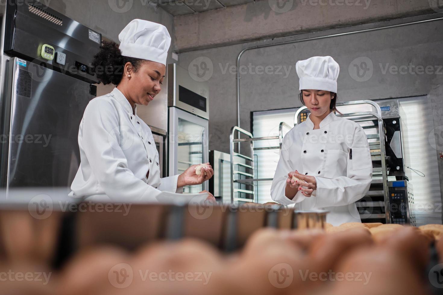 twee professionele vrouwelijke chef-koks in witte kookuniformen en schorten kneden deeg en eieren, bereiden brood, koekjes en vers gebak, bakken in de oven in een roestvrijstalen keuken van een restaurant. foto