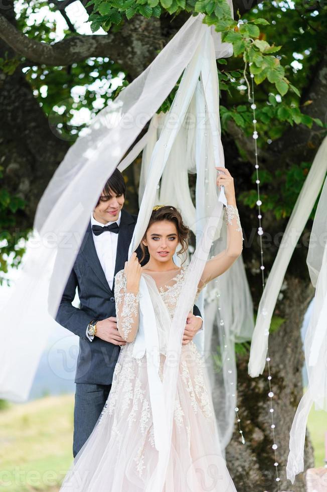 de bruid en bruidegom knuffelen onder een oude eik. bruiloft fotoshoot in de bergen. naast hen is het decor voor de ceremonie voorbereid. foto