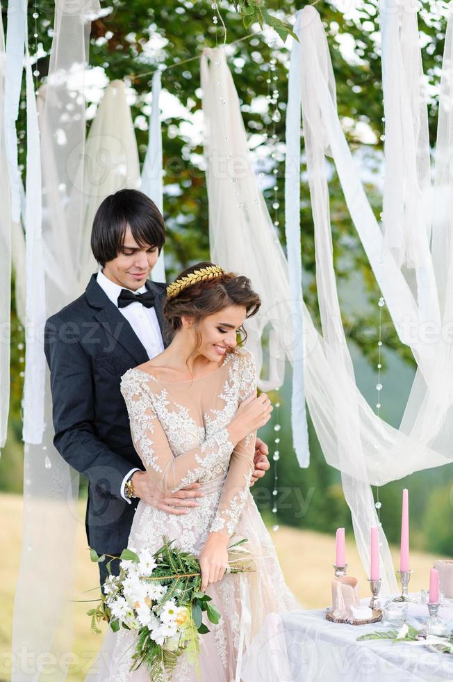 de bruid en bruidegom knuffelen onder een oude eik. bruiloft fotoshoot in de bergen. naast hen is het decor voor de ceremonie voorbereid. foto