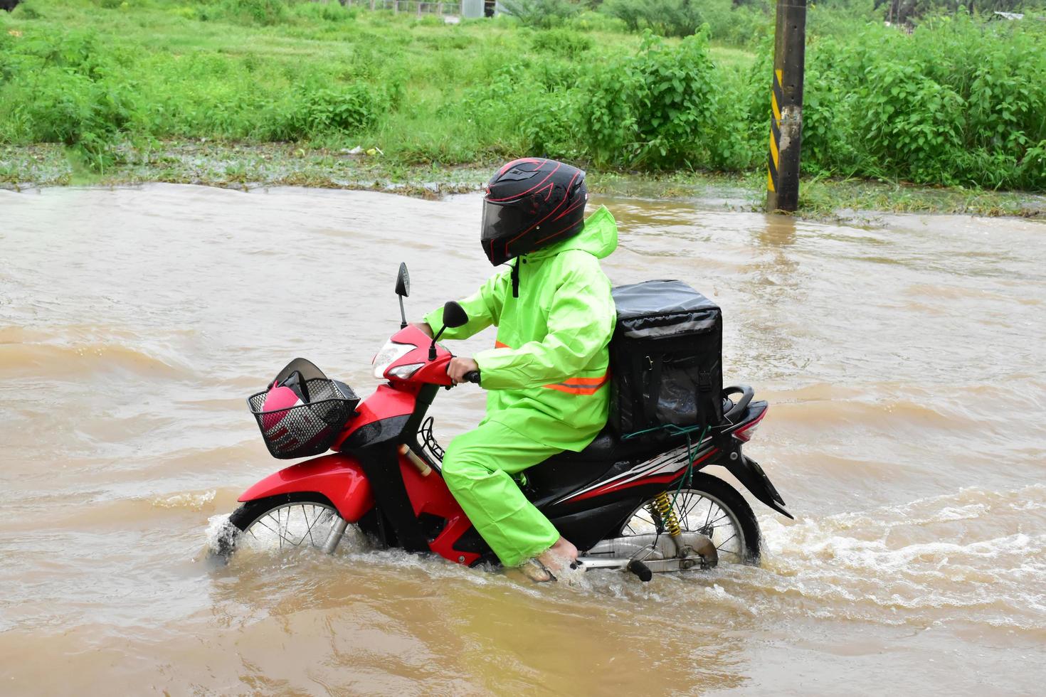 bezorging aziatische man met groen uniform, rijdend en etend bezorgend aan klant met voedselkoffer achter in overstromingsstraat, expresbezorging van voedsel en online winkelen concept in de overstromingssituatie. foto