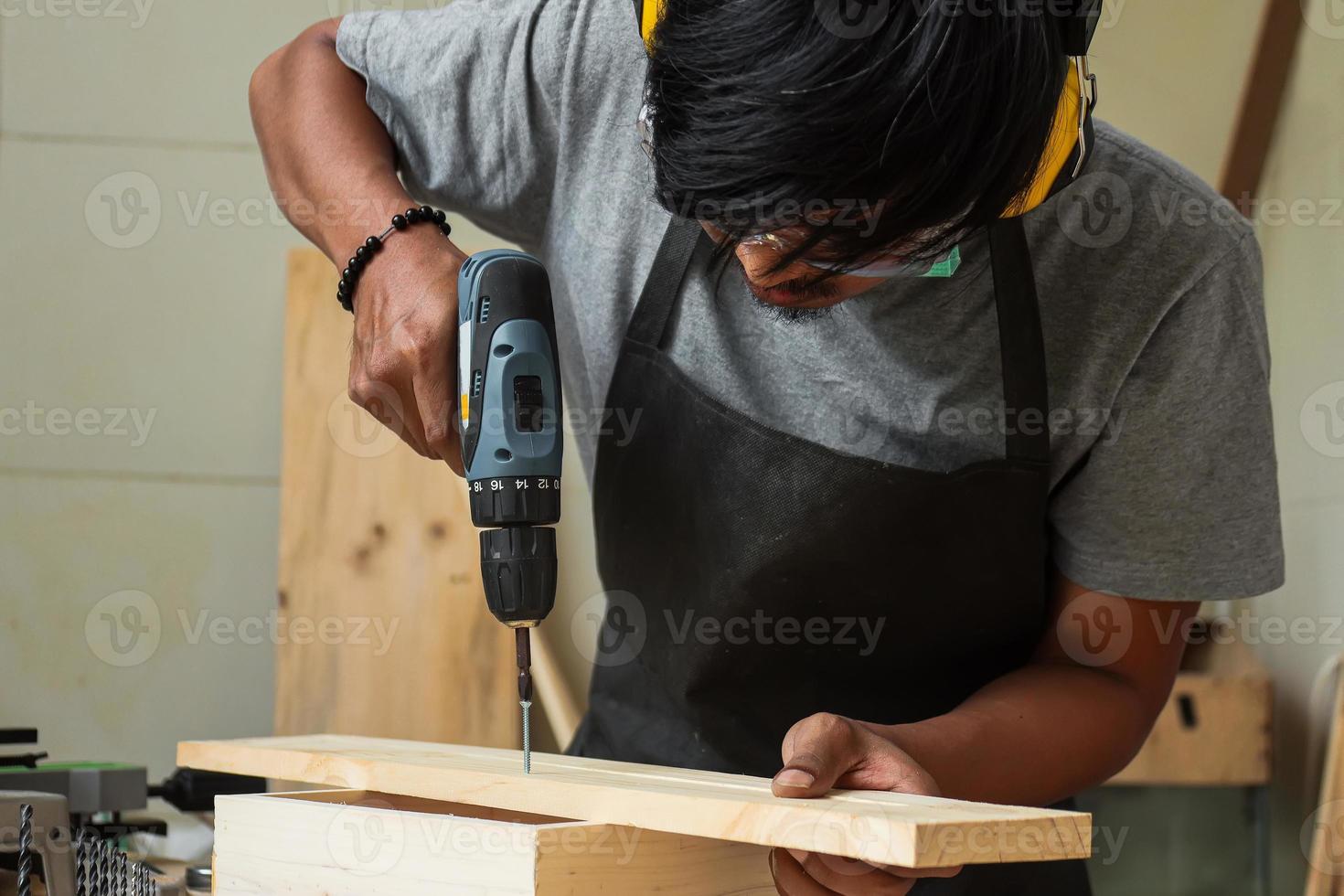 een jonge mannelijke timmerman die aan zijn werkplaatstafel werkt met een boormachine en veiligheidsuitrusting draagt foto