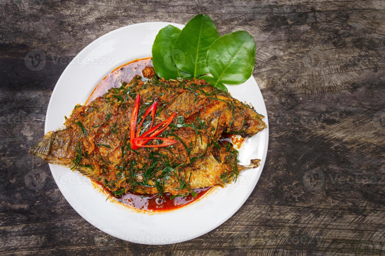 bovenaanzicht gebakken nijl tilapia vis met rode currypasta op witte plaat houten achtergrond, thais eten is pittig vermengd met zoete smaak met aromatische kruiden, rode currypasta is populair om te koken in thailand foto