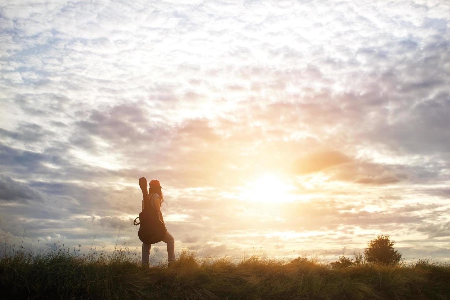 vrouw met een gitaar op haar schouder, plattelandsweg in de prachtige hemel van zonsondergangachtergrond foto