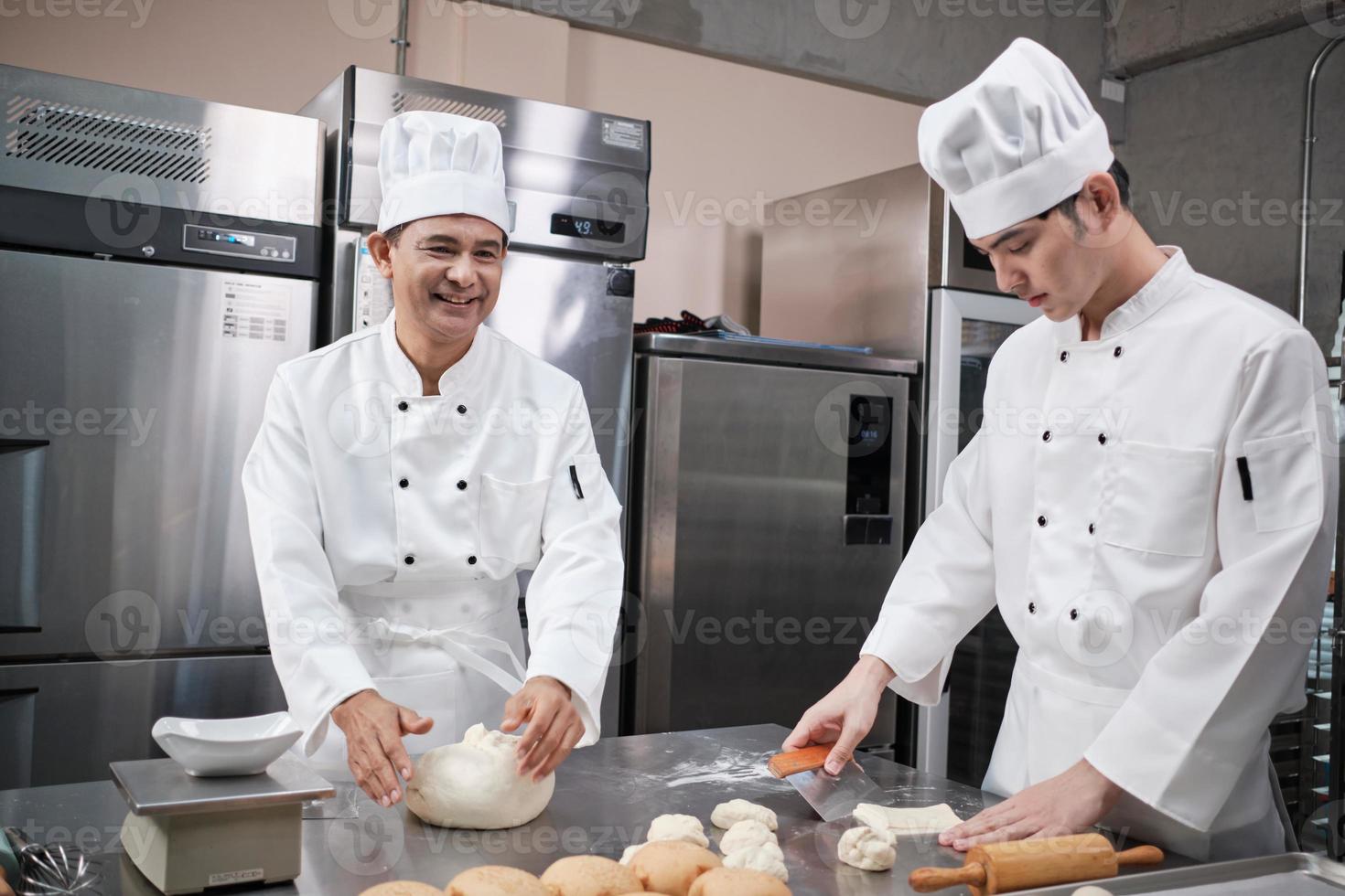twee professionele aziatische mannelijke chef-koks in witte koksuniformen en schorten kneden gebakdeeg en eieren, bereiden brood en vers bakkerijvoedsel, bakken in de oven in de roestvrijstalen keuken van het restaurant. foto