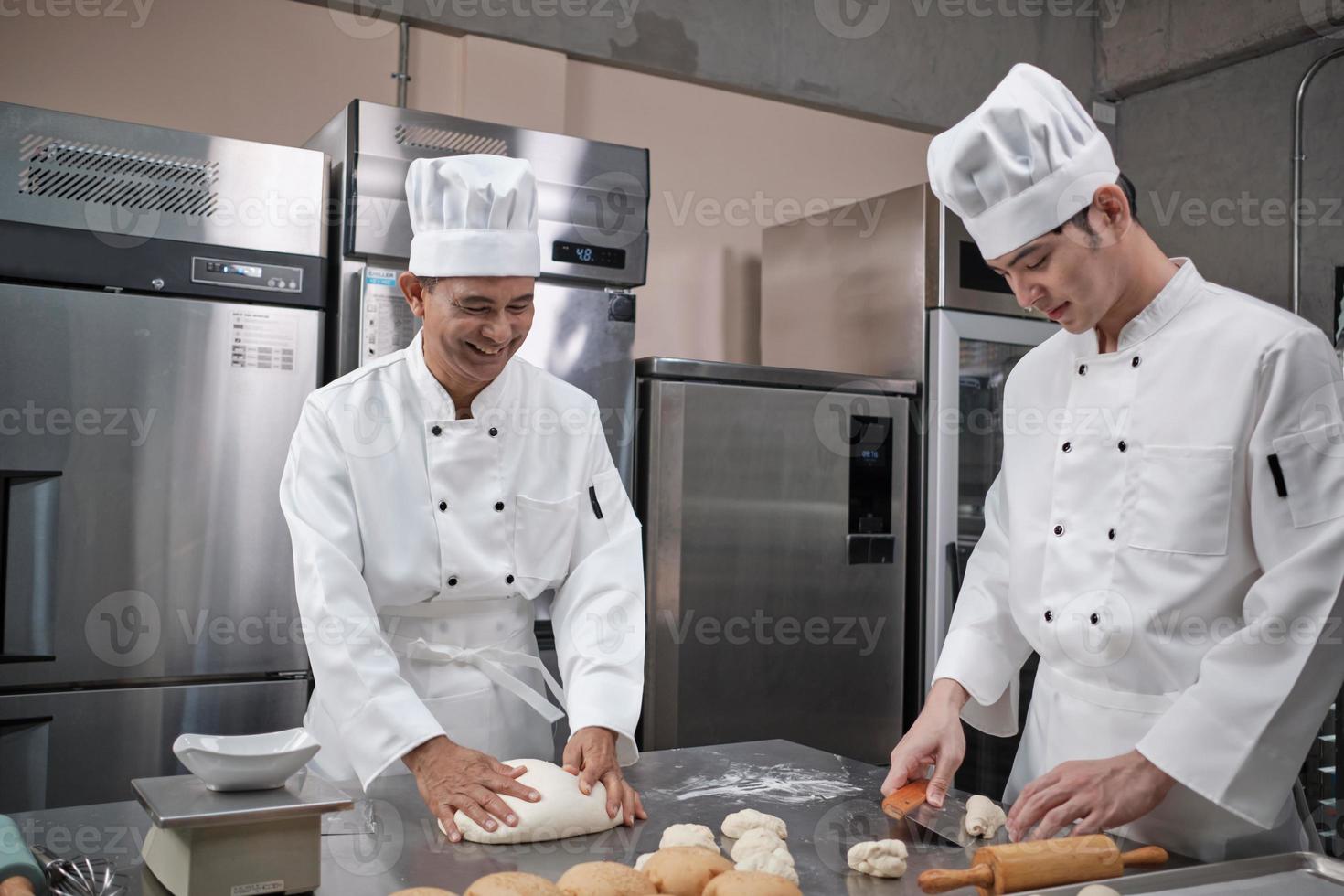 twee professionele aziatische mannelijke chef-koks in witte koksuniformen en schorten kneden gebakdeeg en eieren, bereiden brood en vers bakkerijvoedsel, bakken in de oven in de roestvrijstalen keuken van het restaurant. foto