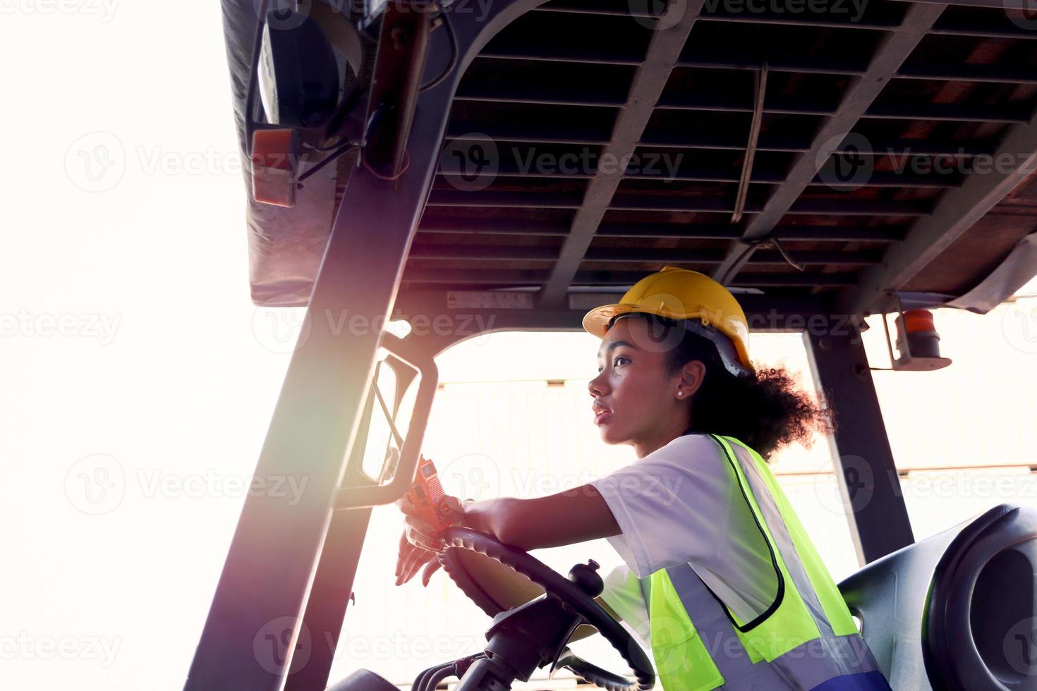 industriële werknemer vrouw dragen veiligheidsvest en helm rijden vorkheftruck auto in de fabrieksindustrie, Afro-Amerikaanse ingenieur mooie vrouw werken bij logistieke verzending vrachtcontainer werf. foto