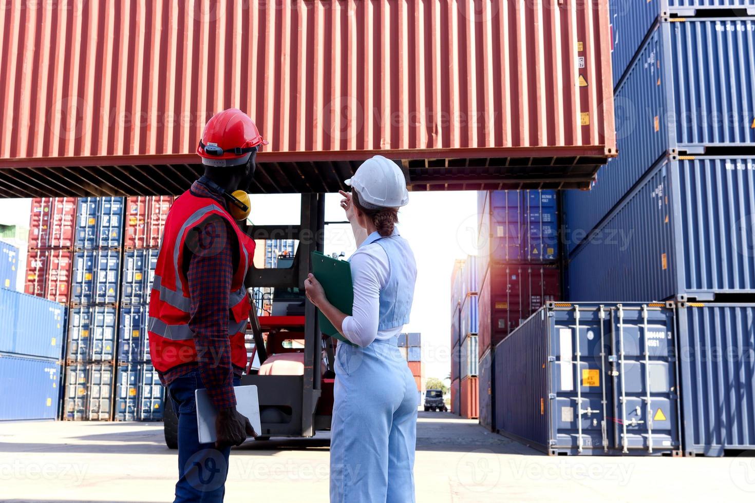 achter twee arbeiders dragen een veiligheidsvest en helm die discussiëren op de logistieke vrachtcontainerwerf. Afro-Amerikaanse ingenieur man praten met mooie jonge vrouw baas met blond haar op de werkplek. foto