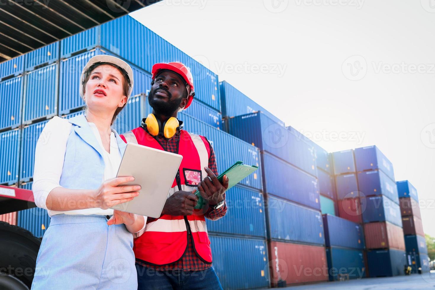 twee arbeiders die een veiligheidsvest en een helm dragen die bij de logistieke containerwerf van de verschepende lading bespreken. Afro-Amerikaanse ingenieur man praten met mooie jonge vrouw baas met blond haar op de werkplek. foto