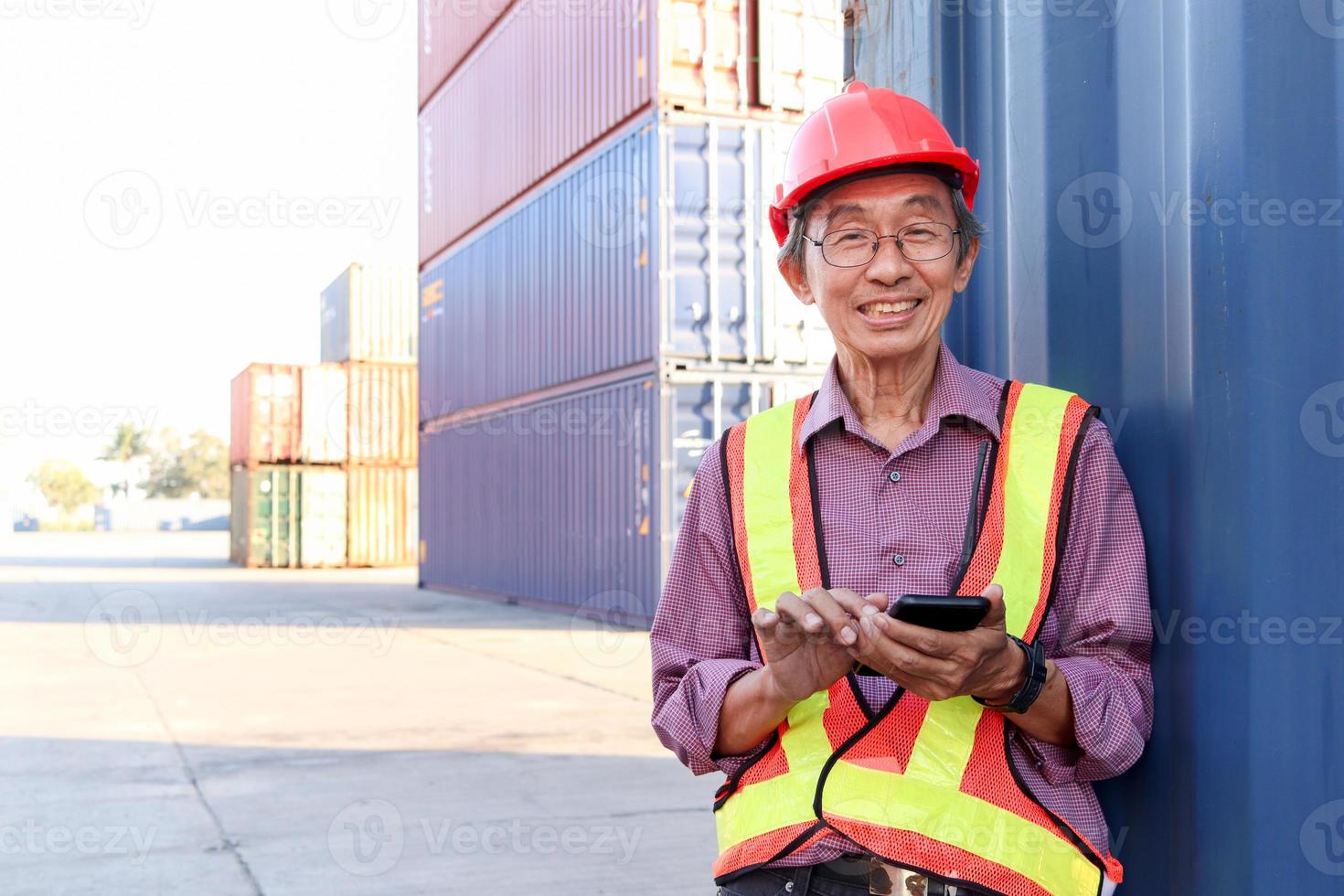 portret van een gelukkig lachende senior oudere aziatische werknemer ingenieur die een veiligheidsvest en helm draagt, staande en mobiele telefoon gebruikt op de werf van logistieke vrachtcontainers. foto