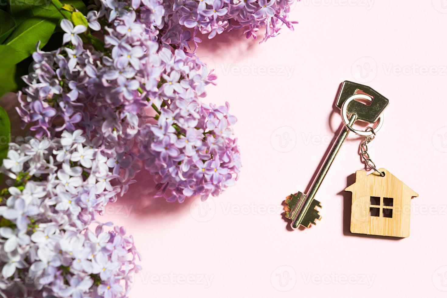 huissleutel met sleutelhanger op roze lente achtergrond en lila takken. zomervakantie huis, huisje op het platteland boeken, verhuizen naar nieuw huis, hypotheek, huur en aankoop onroerend goed. kopieer ruimte foto