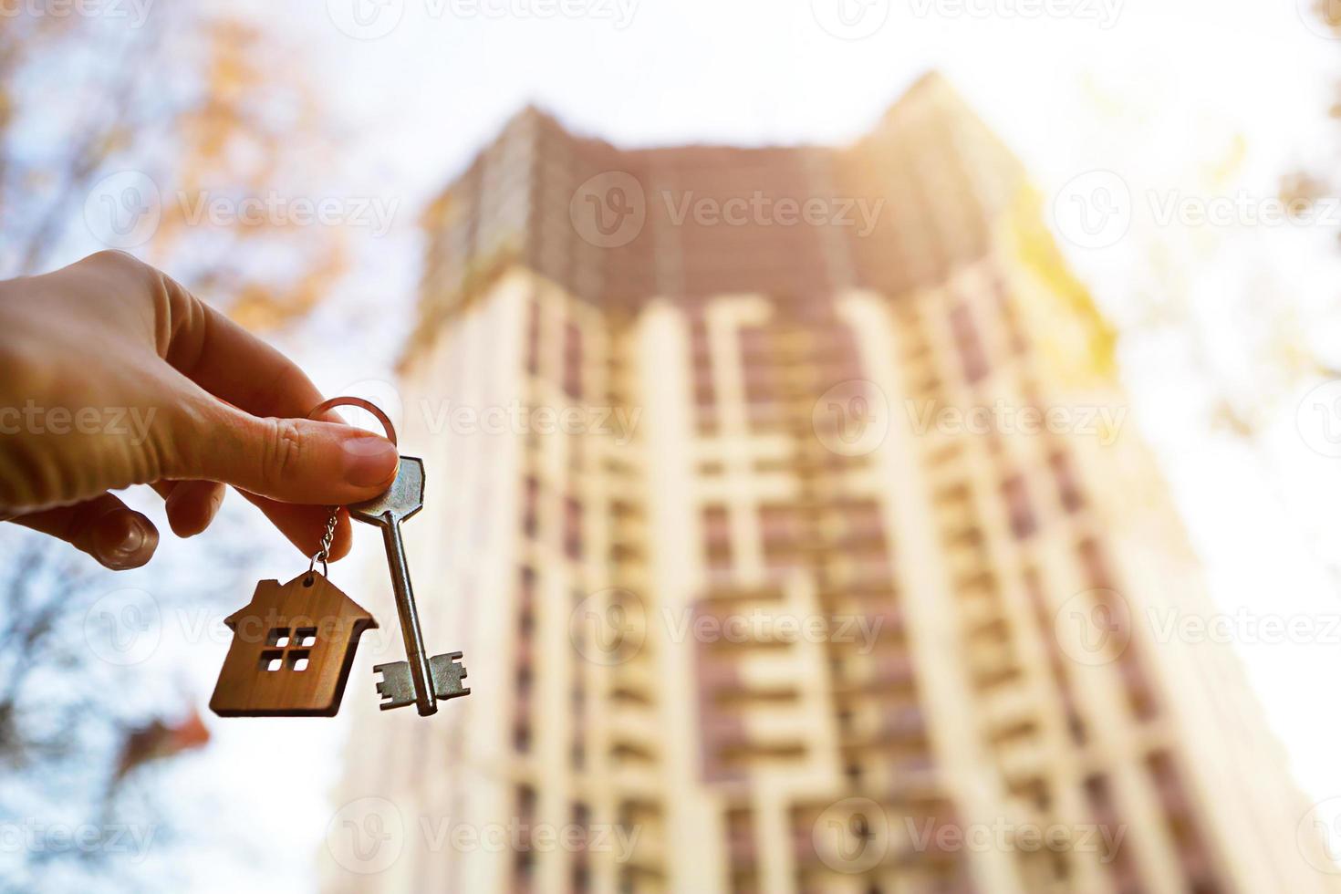 hand met een sleutel en een houten sleutelhanger-huis. achtergrond van wolkenkrabber met meerdere appartementen. bouwen, verhuizen naar een nieuwe flat, hypotheek, huur en aankoop onroerend goed. om de deur te openen. kopieer ruimte foto