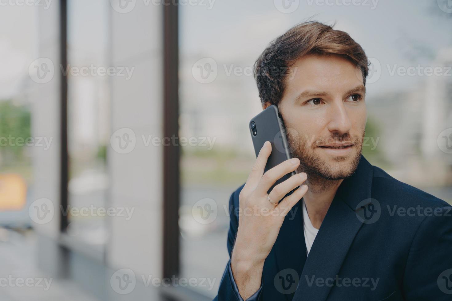 peinzende mannelijke ondernemer gefocust op afstand maakt het beantwoorden van de oproep met behulp van mobiele telefoon foto