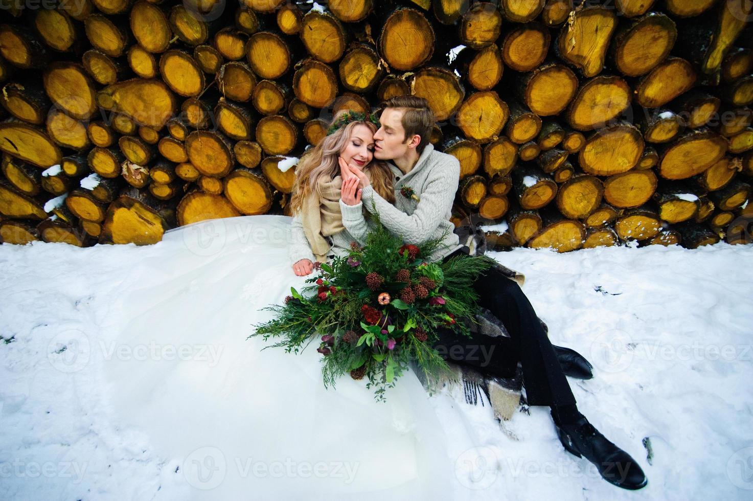bruidegom kust zijn bruid op de tempel. pasgetrouwden met boeket zit op sneeuw op de houten achtergrond. winterhuwelijk foto