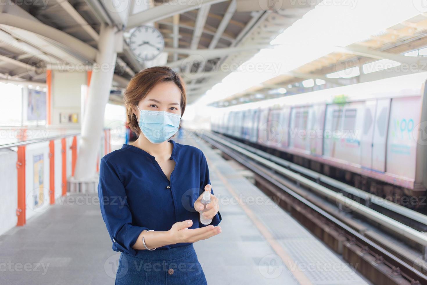 aziatische vrouw wacht op de trein op het station. ze spuit alcohol om haar hand schoon te maken en draagt een medisch gezichtsmasker als gezondheidszorg en een nieuwe normale levensstijl. foto