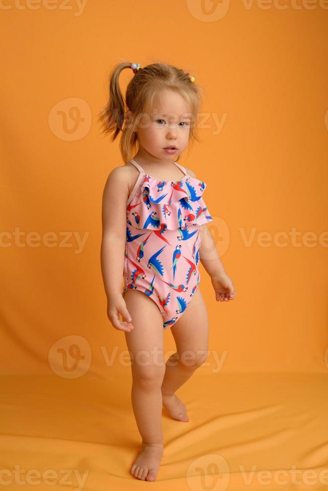 een klein meisje gekleed in een zwempak op de leeftijd van anderhalf jaar springt of danst. het meisje is erg blij. foto genomen in de studio op een gele achtergrond.