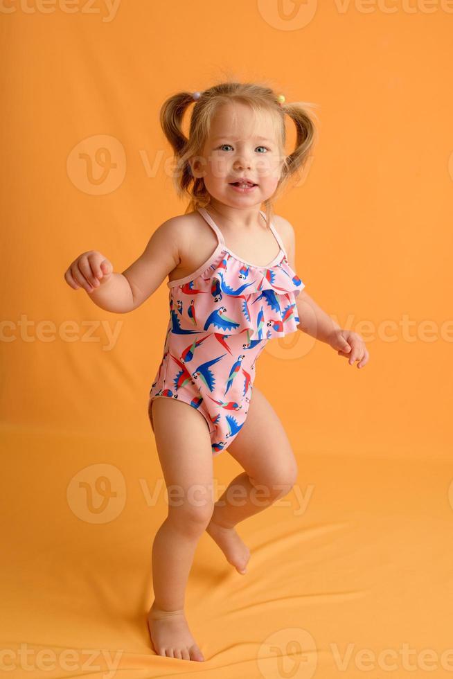 een klein meisje gekleed in een zwempak op de leeftijd van anderhalf jaar springt of danst. het meisje is erg blij. foto genomen in de studio op een gele achtergrond.