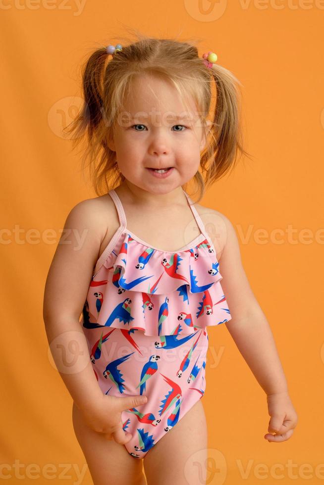 een klein meisje gekleed in een zwempak op de leeftijd van anderhalf jaar springt of danst. het meisje is erg blij. foto genomen in de studio op een gele achtergrond.