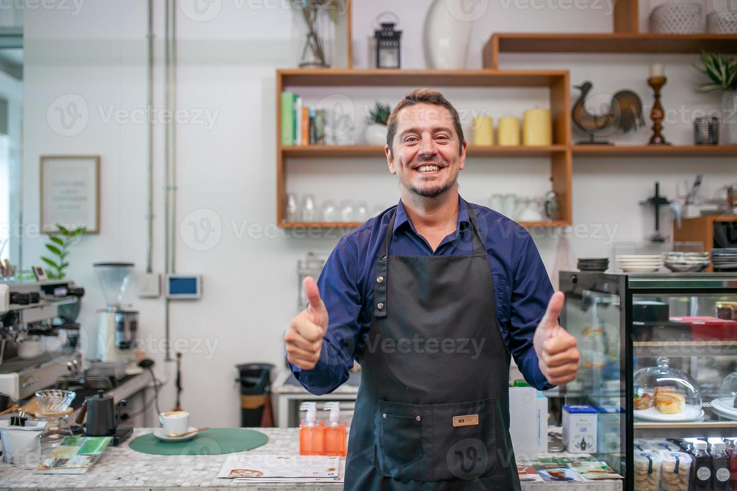 portret van een lachende eigenaar die in zijn café staat. koffie-eigenaar staat met schort in de coffeeshop om de klant te verwelkomen. foto
