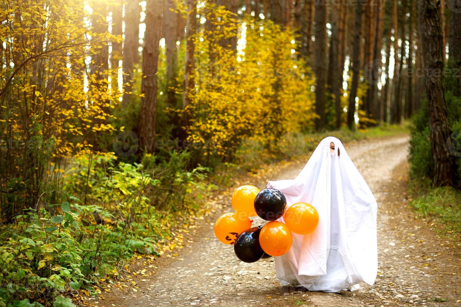 een kind in lakens met spleten als een spookkostuum in een herfstbos met oranje en zwarte ballen schrikt. Halloween feest foto