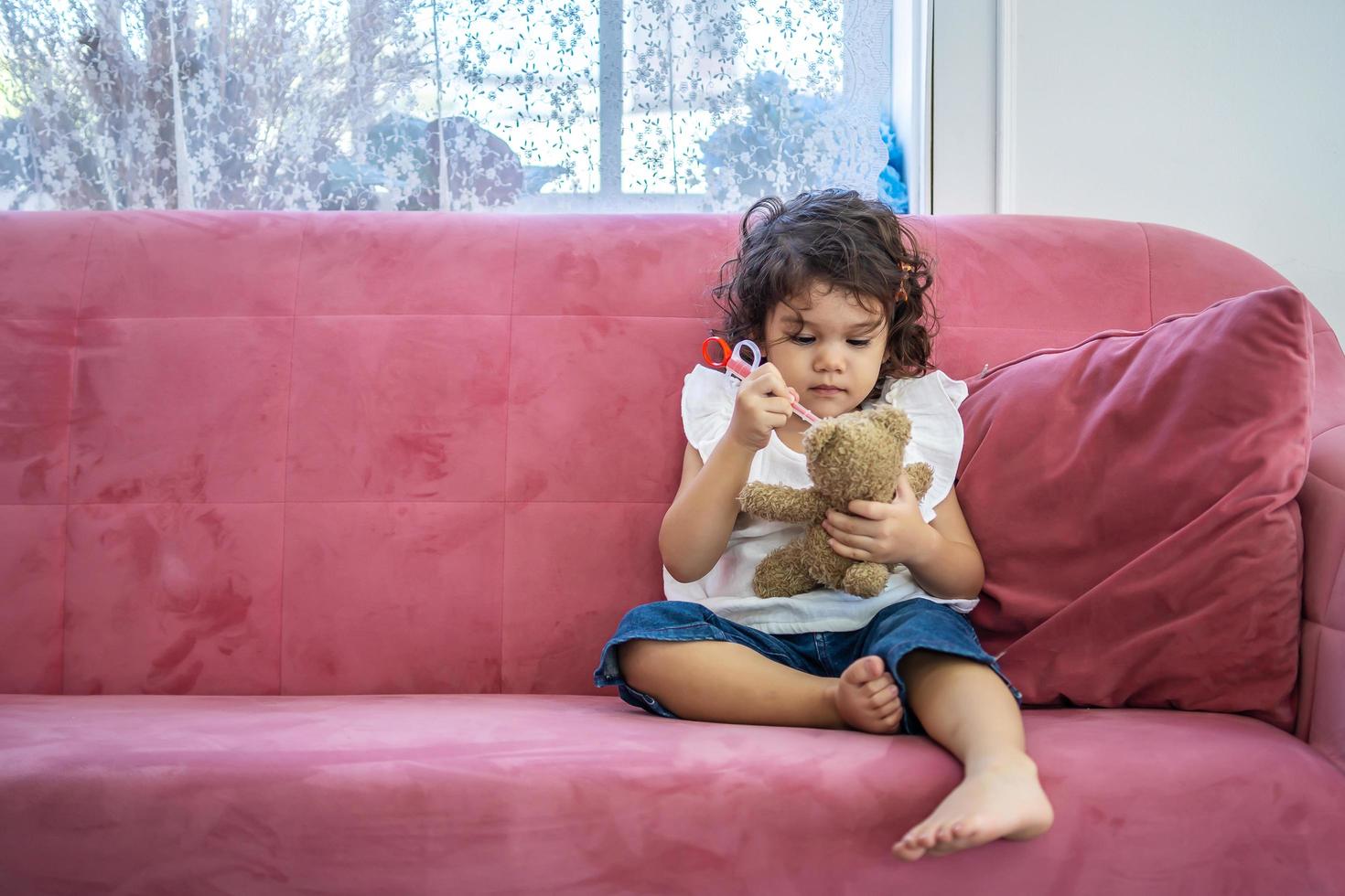 een klein schattig meisje geniet van het spelen van speelgoed injectiespuiten speelgoed op de teddybeer op de bank in het huis, onderwijstijd thuis foto