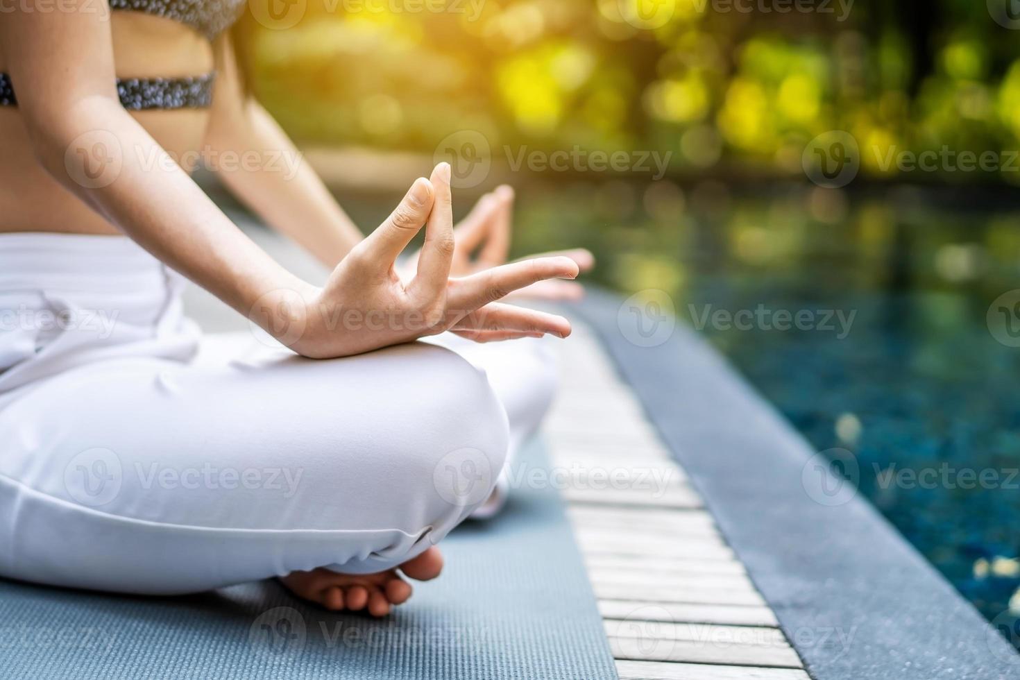 close-up van haar handen en gekruiste benen in de lotuspositie, vrouw die yoga en meditatie traint in een lotusyoga-positie bij blauw zwembad foto