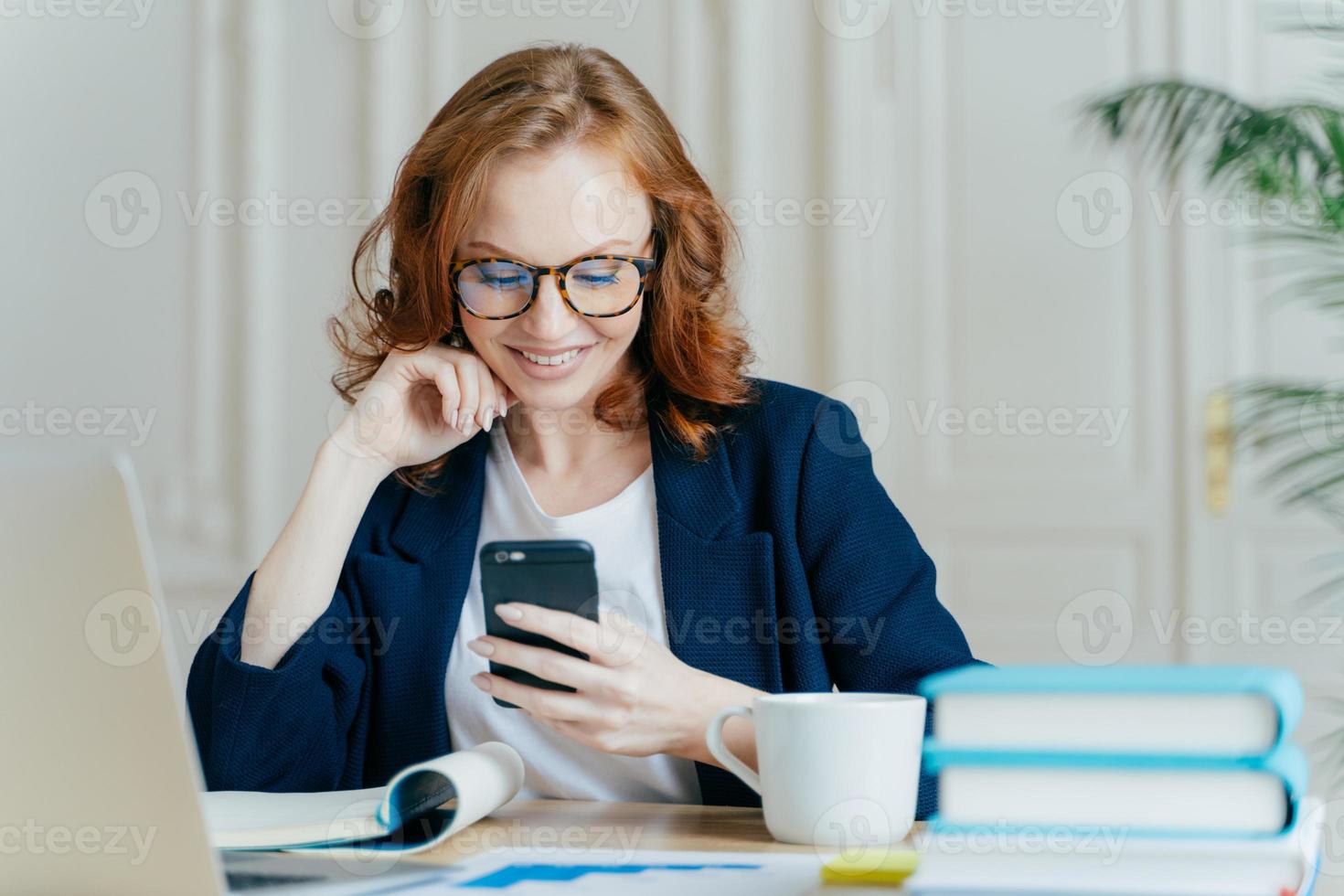 foto van mooie vrouw zit met smartphoneapparaat, typt feedback, werkt op kantoor op up-to-date laptop, gefocust op scherm van gadget, zit op werkplek met boeken, notitieblok en warme drank