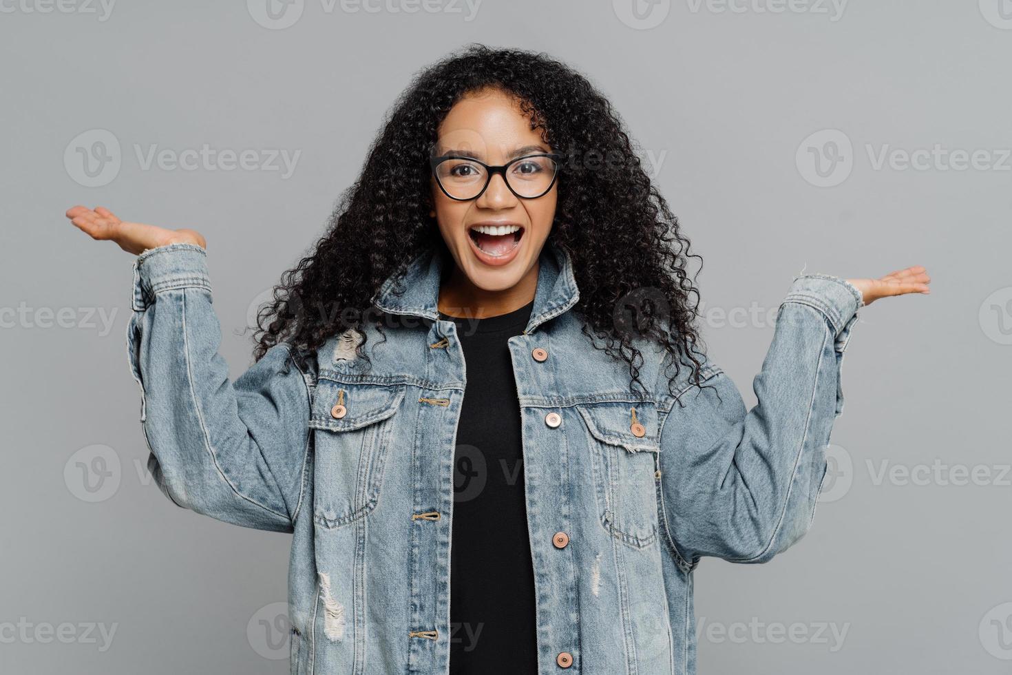 portret van blij tevreden krullend Afro-Amerikaanse vrouw heft handpalmen op, doet alsof ze iets vasthoudt, draagt een optische bril en een spijkerjasje, geïsoleerd op een grijze achtergrond. mensen en geluk concept foto