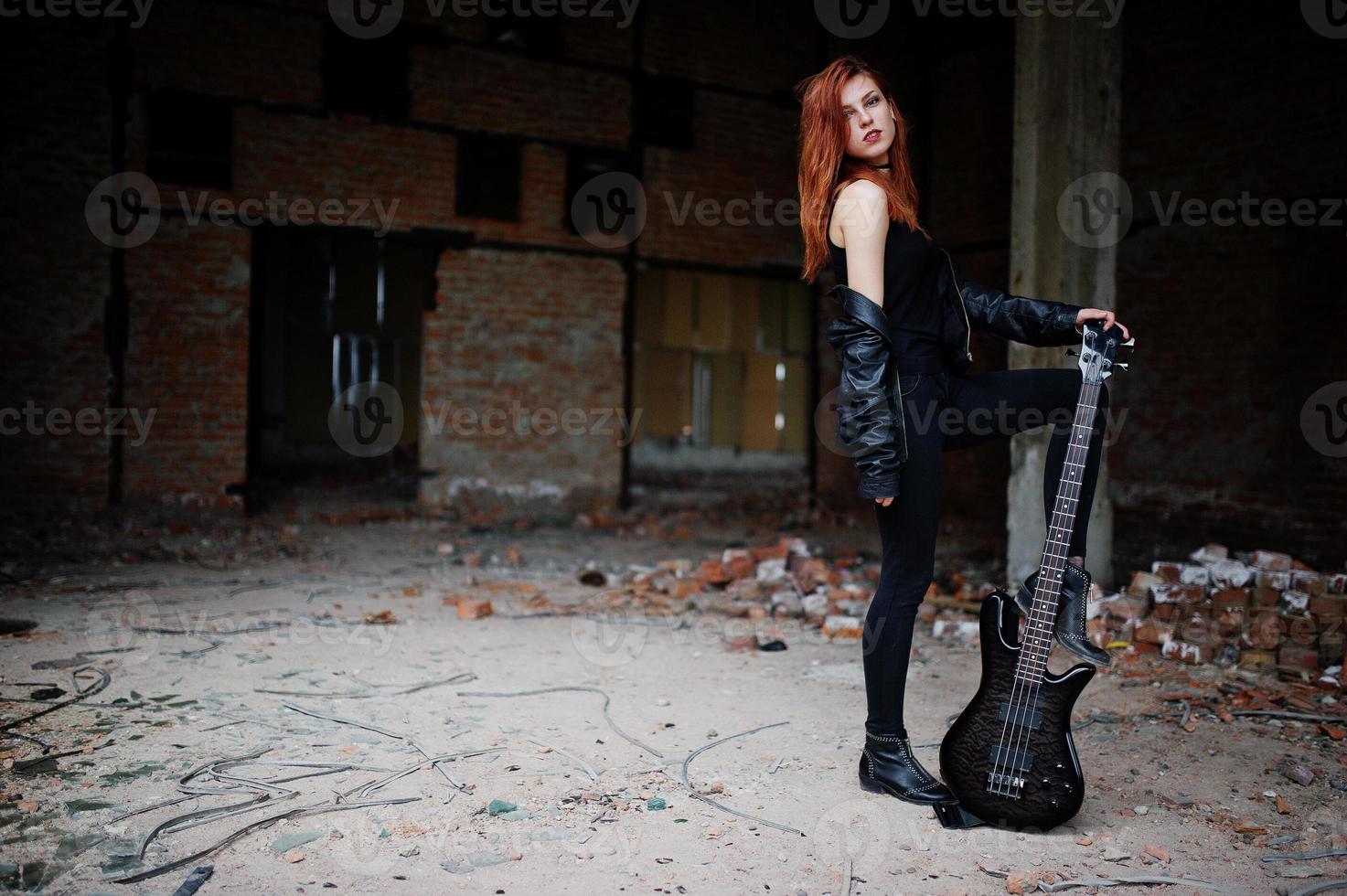 roodharige punkmeisjeslijtage op zwart met basgitaar op verlaten plaats. portret van gotische vrouw muzikant. foto