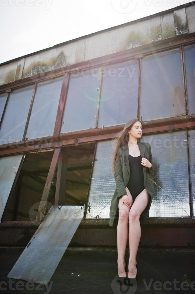 sexy model meisje wit lange benen bij zwarte lingerie outfit body badpak combidress en jas gesteld op het dak van verlaten industriële plaats met ramen. foto
