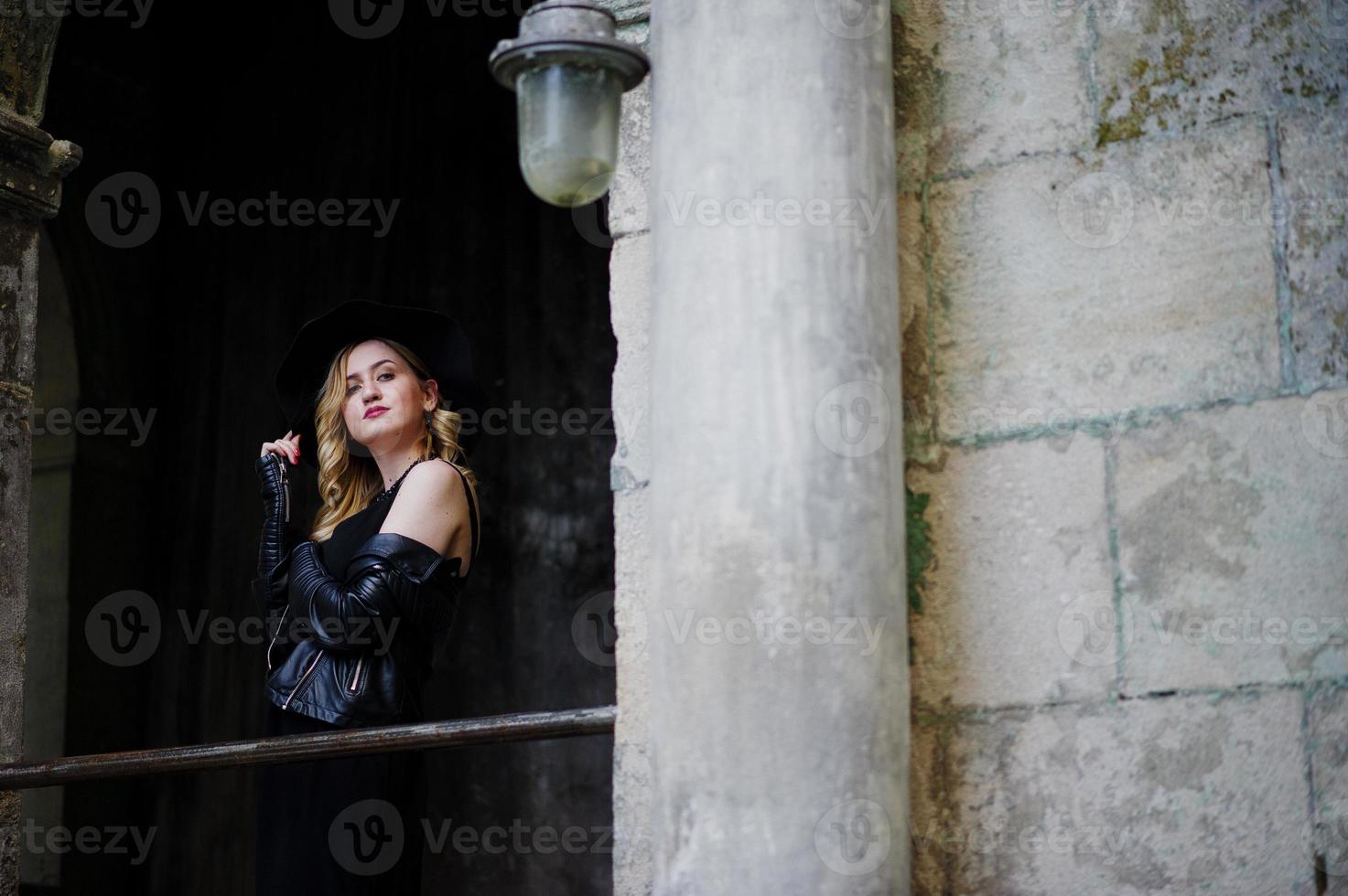 blonde vrouw op zwarte jurk, leren jas, kettingen en hoed tegen de oude straat van de stad. foto