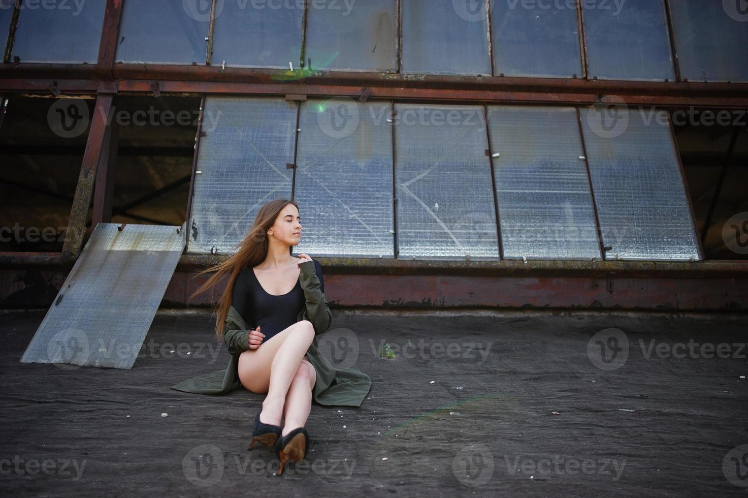 sexy model meisje wit lange benen bij zwarte lingerie outfit body badpak combidress en jas gesteld op het dak van verlaten industriële plaats met ramen. foto