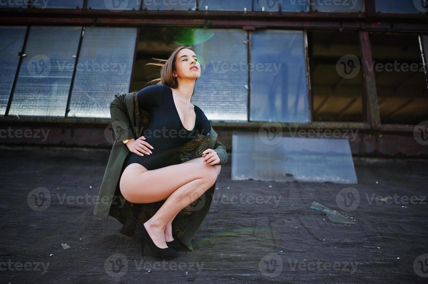 sexy model meisje wit lange benen bij zwarte lingerie outfit body badpak combidress en jas gesteld op het dak van verlaten industriële plaats met ramen. foto