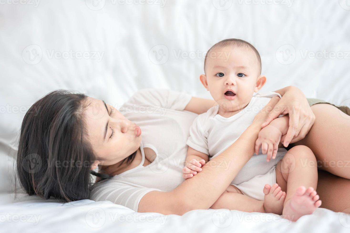 schattige Aziatische pasgeboren babyjongen zit spelen op wit bed met een blij gezicht van de glimlach. terwijl je moeder in de buurt zorgt. kleine onschuldige nieuwe baby schattig kind in de eerste dag van het leven. moederdagconcept. foto