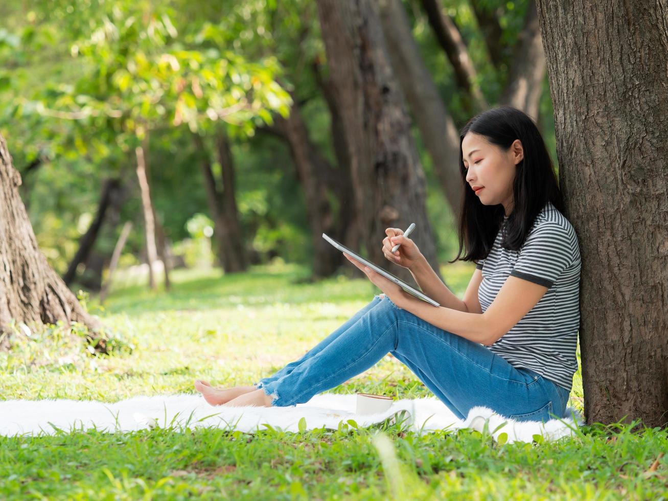 een mooie Aziatische vrouw zit in een tuin en schrijft informatie van internet op een digitale tablet foto