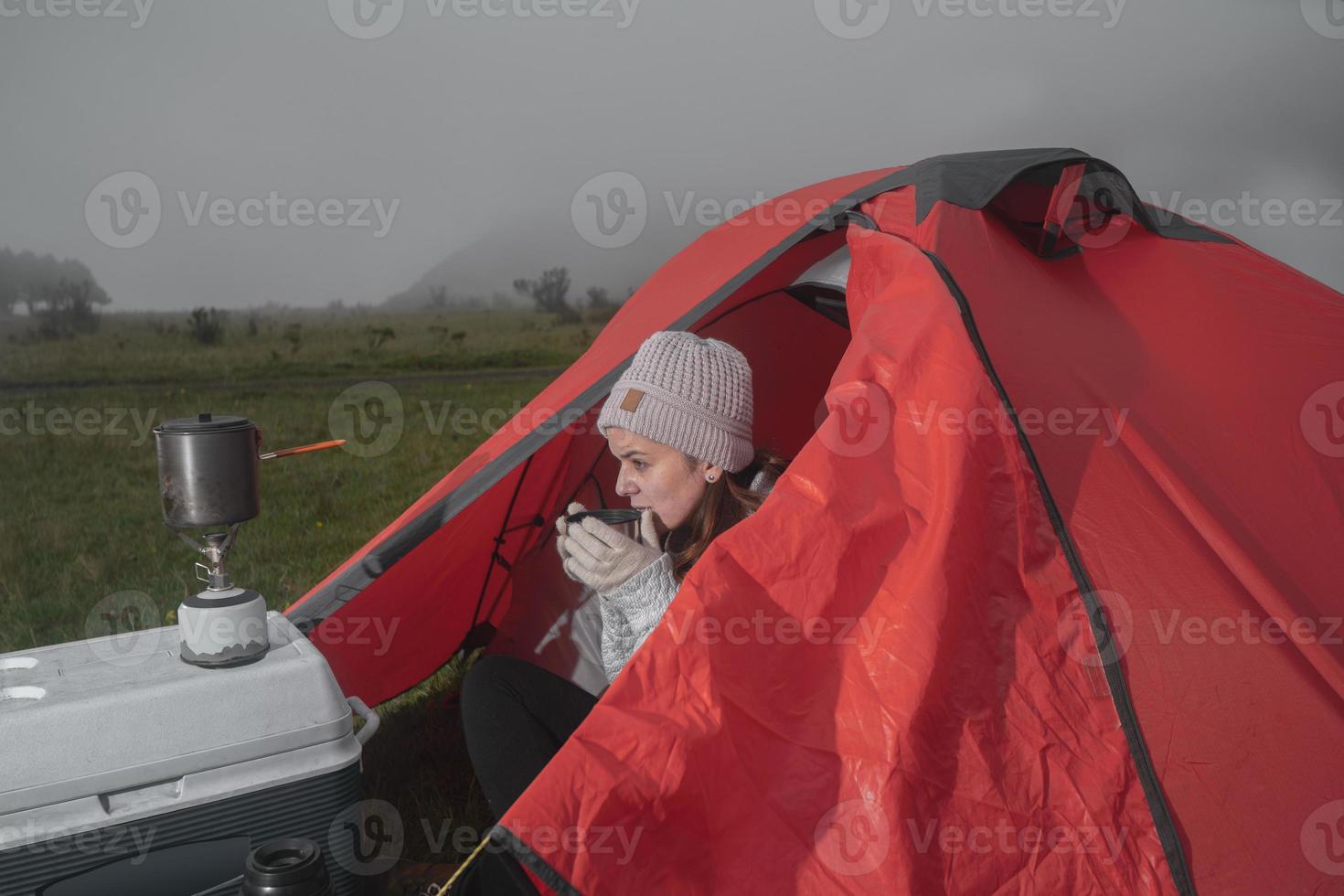 mooie latijnse vrouw die buiten haar tent zit en thee drinkt uit haar thermos op een bewolkte zonsopgang foto