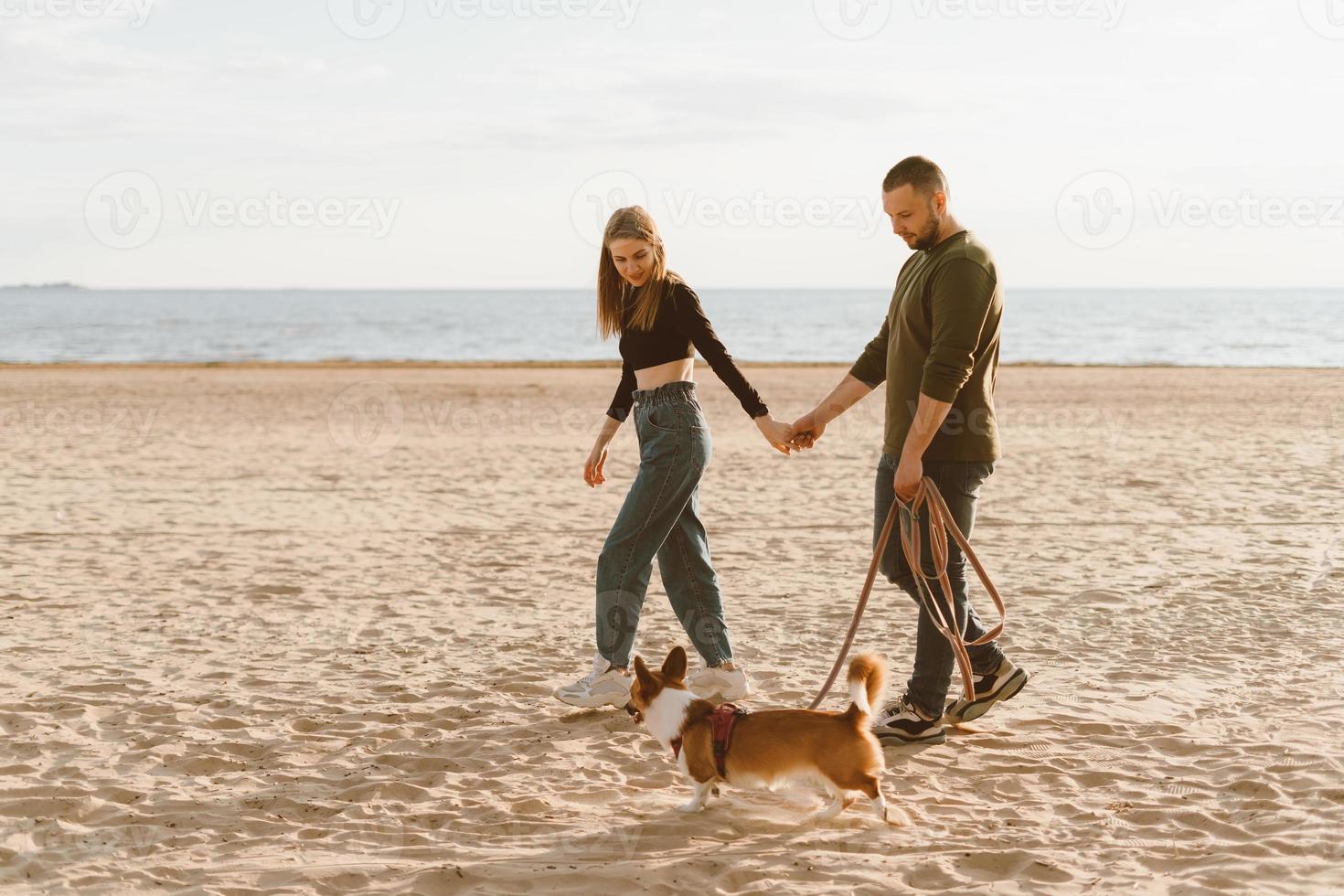 jonge gelukkige paar en hond wandelen langs het strand. man met hand van vrouw en corgi puppy foto