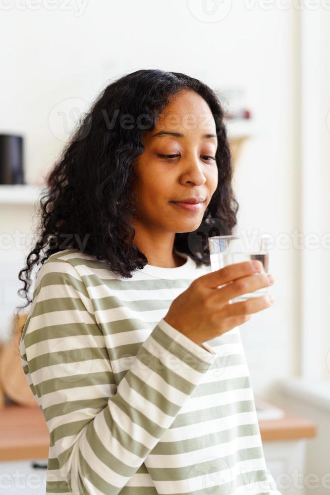 lachende Afro-Amerikaanse vrouw kijkt naar glas helder water op zachte gerichte achtergrond foto